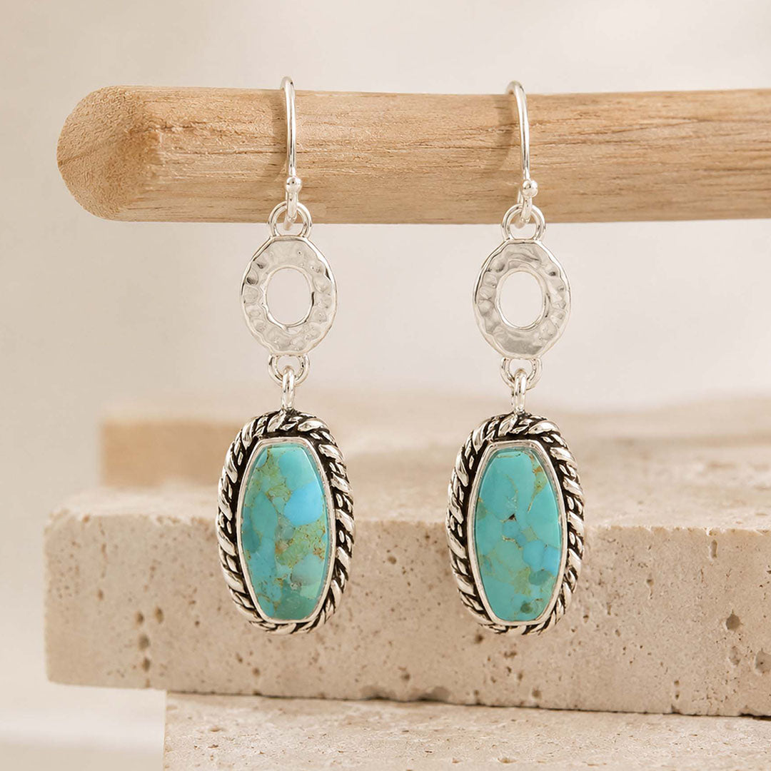 A pair of silver dangle earrings with oval turquoise stones in braided silver bezels, hanging from a wooden dowel against a neutral beige background.