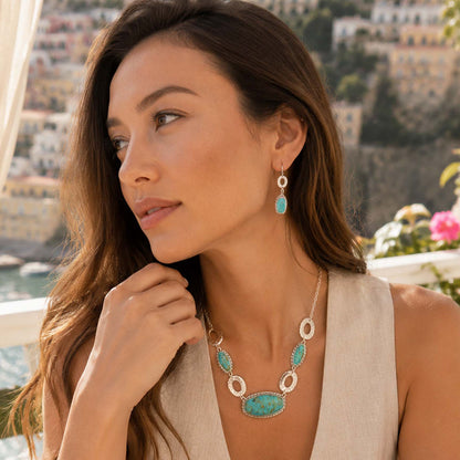 A woman wearing a turquoise and silver statement necklace and matching drop earrings, photographed outdoors with a coastal town in the background.