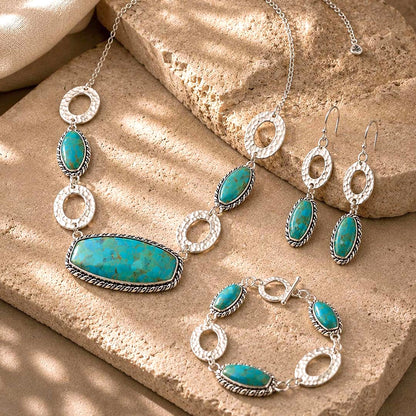 A jewelry set featuring a necklace, pair of drop earrings, and a bracelet, all in silver with turquoise-colored oval and elongated stones, arranged on textured tan stone blocks.