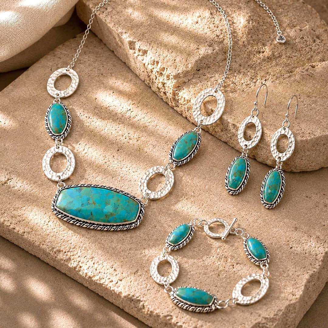 A jewelry set featuring a necklace, pair of drop earrings, and a bracelet, all in silver with turquoise-colored oval and elongated stones, arranged on textured tan stone blocks.