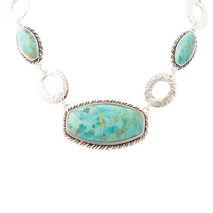 A sterling silver necklace featuring three oval turquoise stones and two textured circular links, with a larger central rectangular turquoise stone, set against a plain white background.