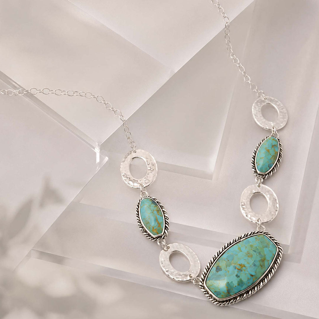 A silver-toned necklace featuring three oval turquoise stones and textured silver rings, displayed on a clean white geometric surface.