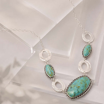 A silver-toned necklace featuring three oval turquoise stones and textured silver rings, displayed on a clean white geometric surface.