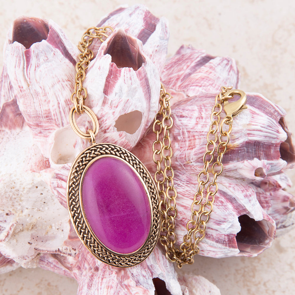 An oval pendant necklace with a vibrant magenta-purple cabochon stone and a gold-toned braided bezel and chain, displayed on a pinkish seashell.
