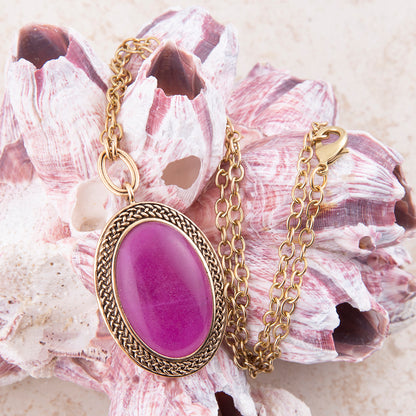 An oval pendant necklace with a vibrant magenta-purple cabochon stone and a gold-toned braided bezel and chain, displayed on a pinkish seashell.