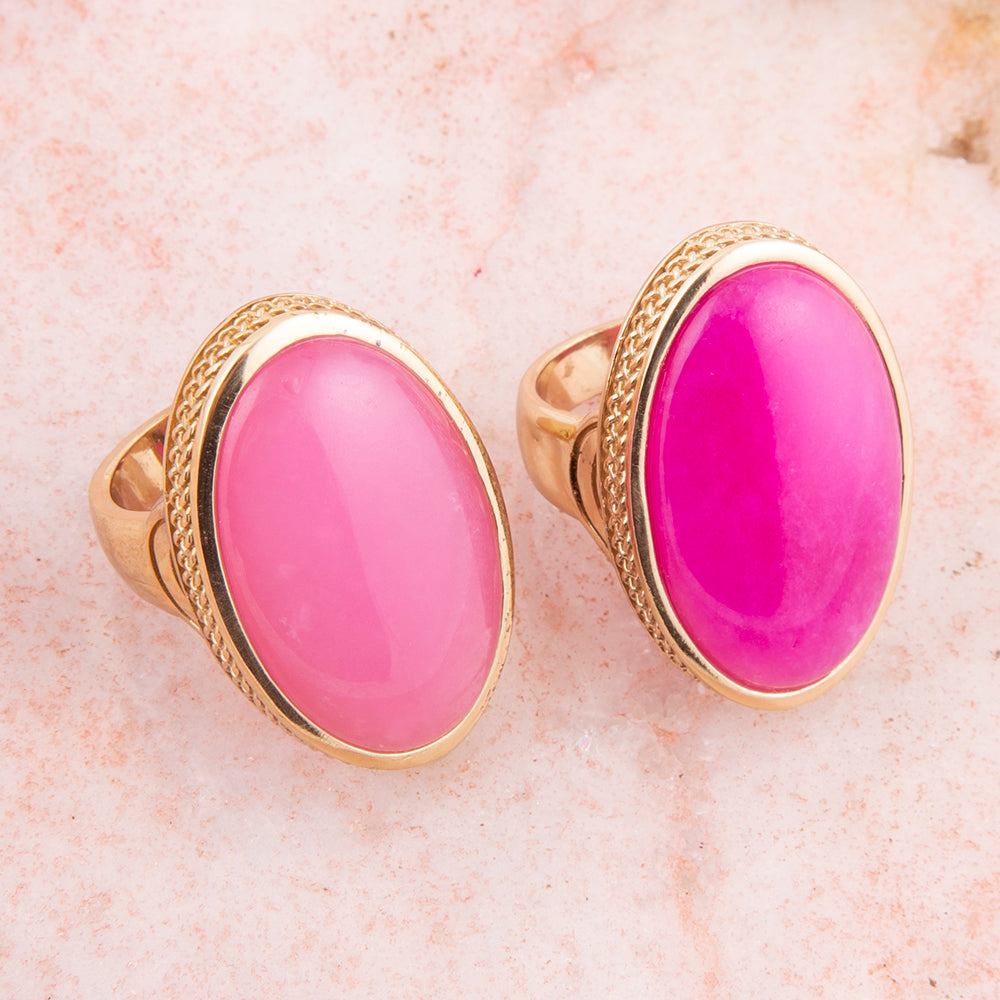 A pair of gold signet rings with oval cabochon gemstones in shades of pink and magenta, displayed on a light pink textured surface.