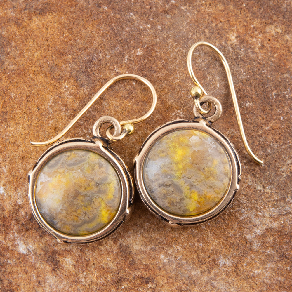 A pair of round drop earrings with textured yellow and gray marbled cabochon stones in silver bezels, hanging from gold-tone fishhook wires on a textured brown stone background.