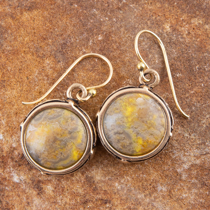A pair of round drop earrings with textured yellow and gray marbled cabochon stones in silver bezels, hanging from gold-tone fishhook wires on a textured brown stone background.