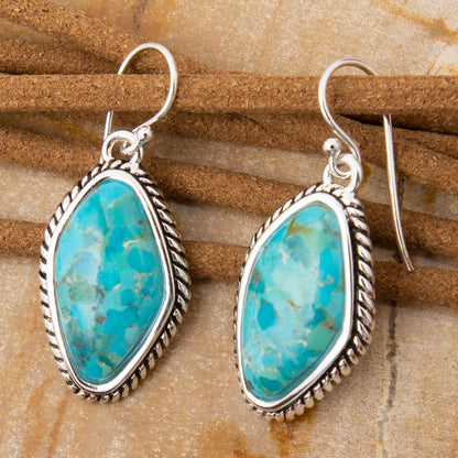 A pair of sterling silver dangle earrings with organic-shaped turquoise stones, displayed on a textured brown leather strap over a tan stone surface.