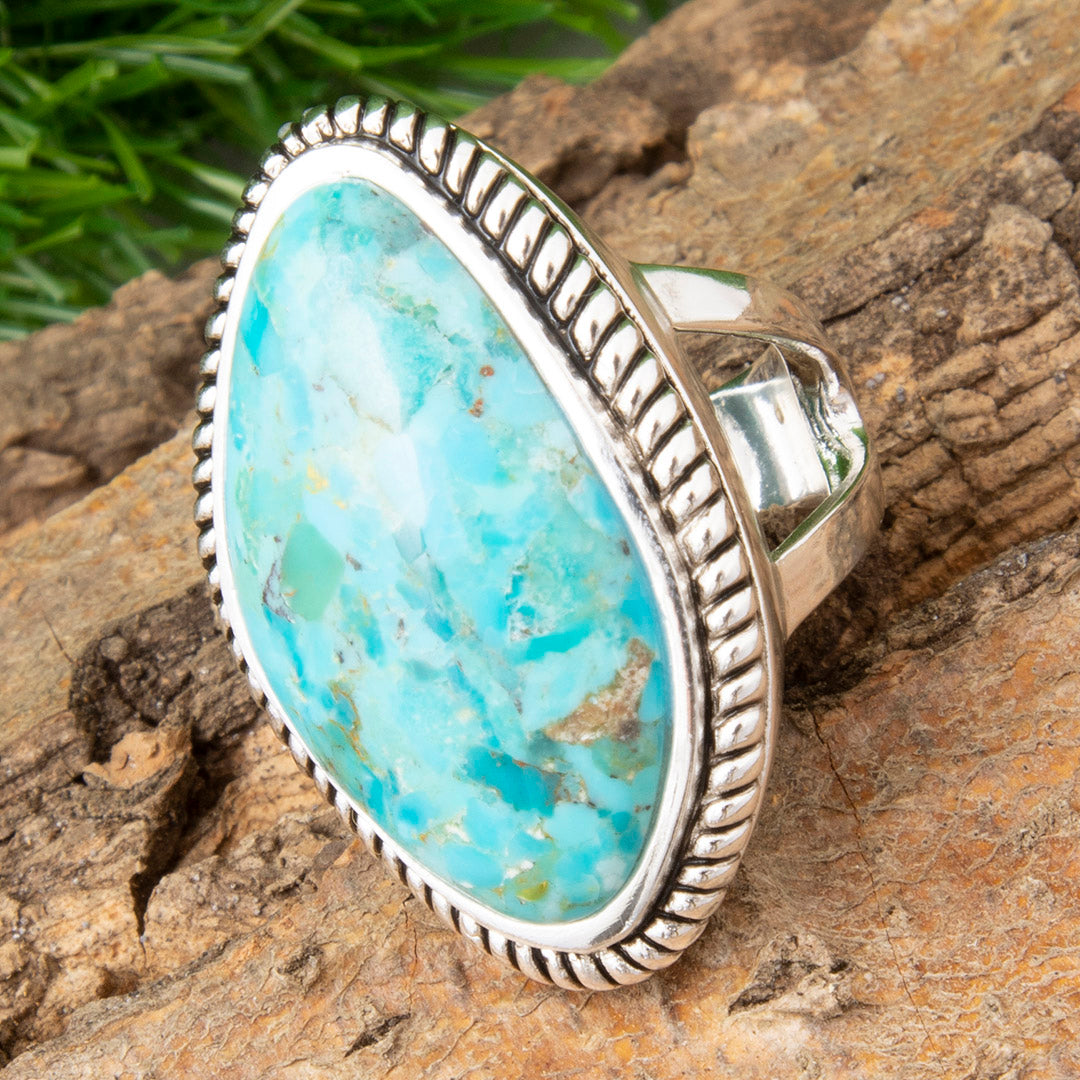 A sterling silver ring featuring a large oval turquoise cabochon with natural white and brown matrix veining, set in a textured beaded silver bezel and resting on a rustic wood surface with green grass in the background.