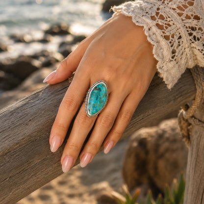 A close-up lifestyle shot of a hand wearing a silver ring with a large oval turquoise stone, resting on a weathered wooden post with a beach background.