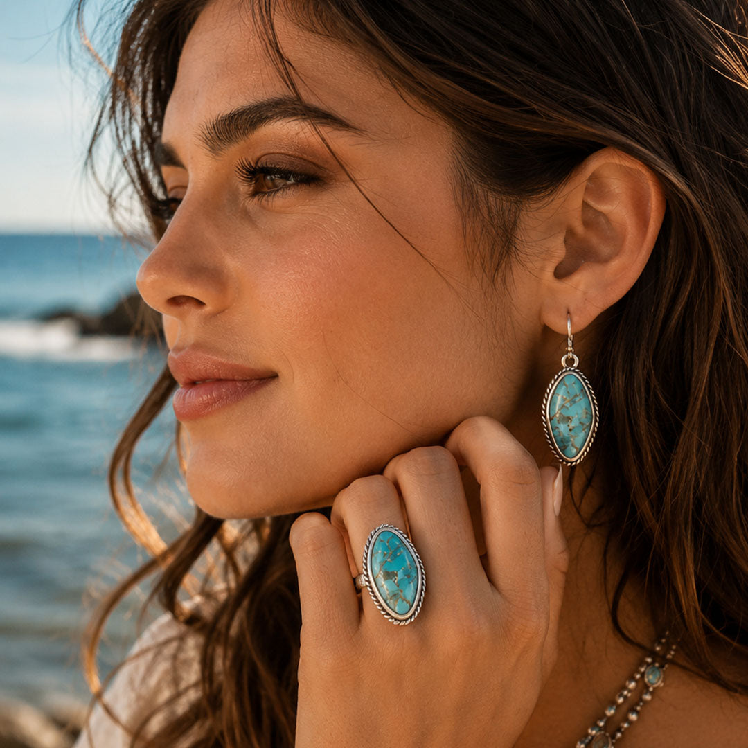 A woman at the beach wearing a silver ring and matching drop earring, both featuring oval turquoise stones with a silver beaded border.