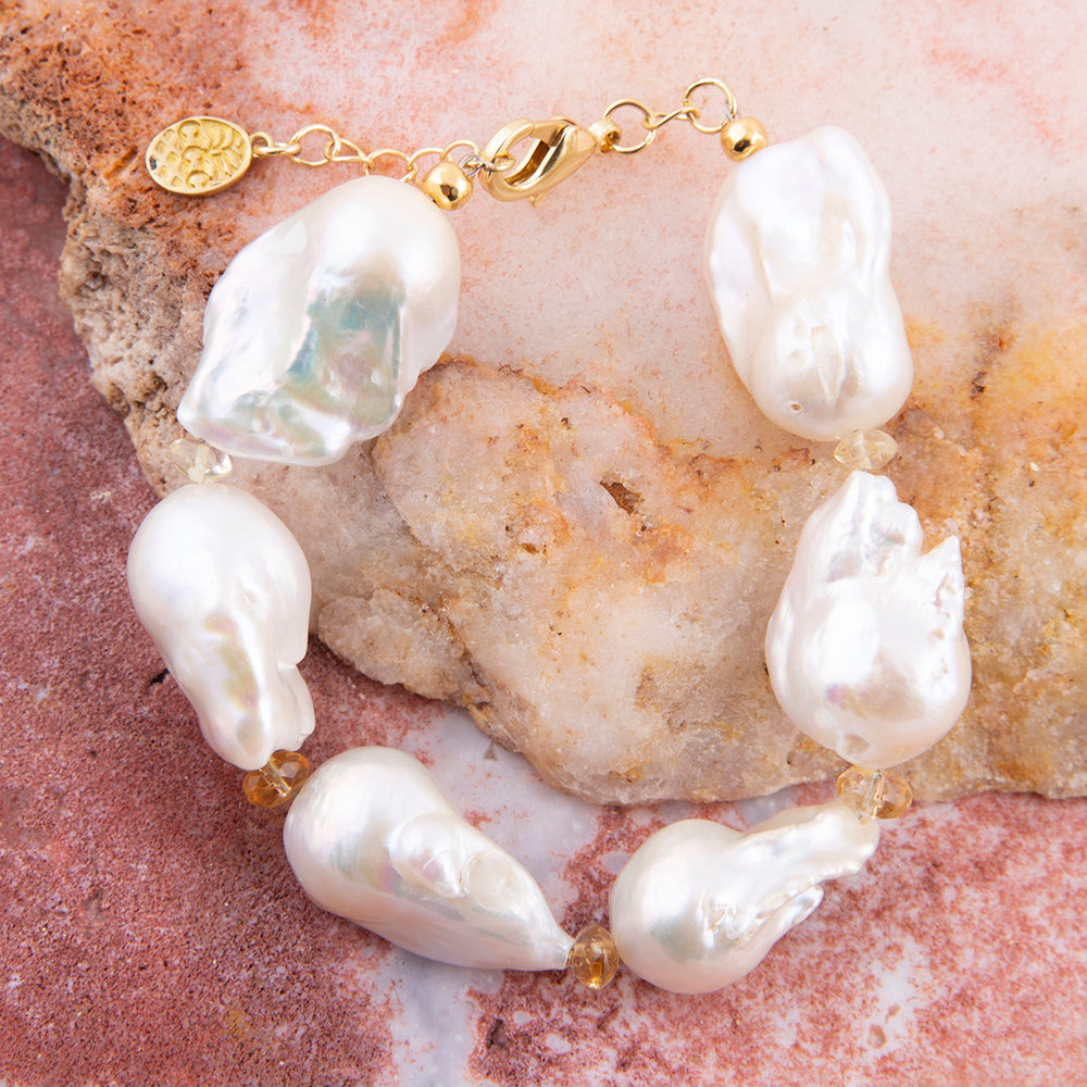 A baroque pearl bracelet with irregular white pearls and small gold spacer beads, displayed on a textured pink and beige stone surface.