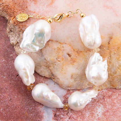 A baroque pearl bracelet with irregular white pearls and small gold spacer beads, displayed on a textured pink and beige stone surface.