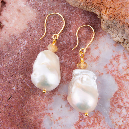 A pair of gold drop earrings with irregular white baroque pearls and small clear beads, displayed on a pinkish stone surface.
