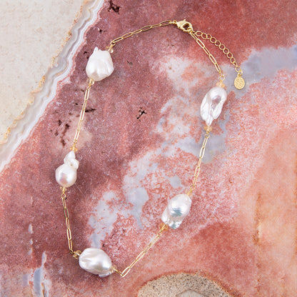 A gold chain necklace featuring irregular white baroque pearls, displayed on a textured pink and beige agate slice background.