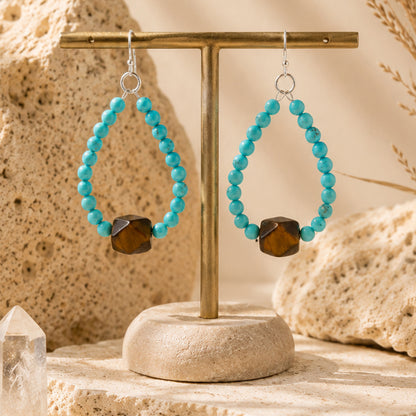 Turquoise beaded hoop earrings with a faceted brown gemstone, displayed on a gold T-stand against a warm, textured stone background.