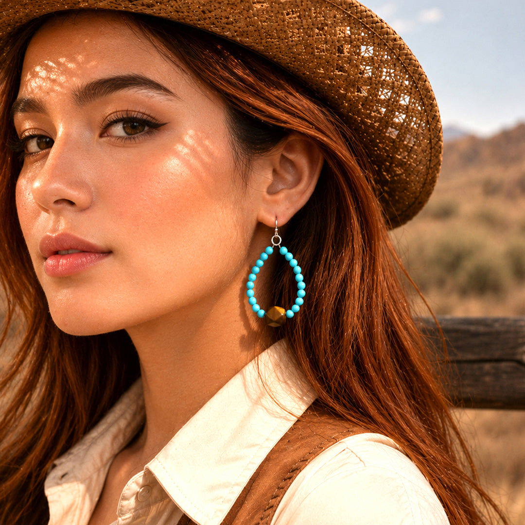 A woman in a brown woven hat and white shirt wearing a pair of large turquoise beaded teardrop earrings, photographed outdoors with warm sunlight and a blurred desert landscape in the background.