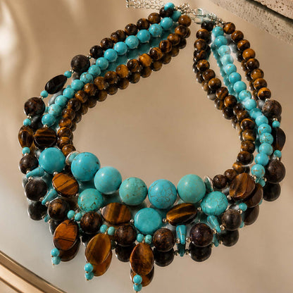 A multi-strand statement necklace featuring turquoise, dark brown, and tiger's eye beads, displayed on a reflective surface.