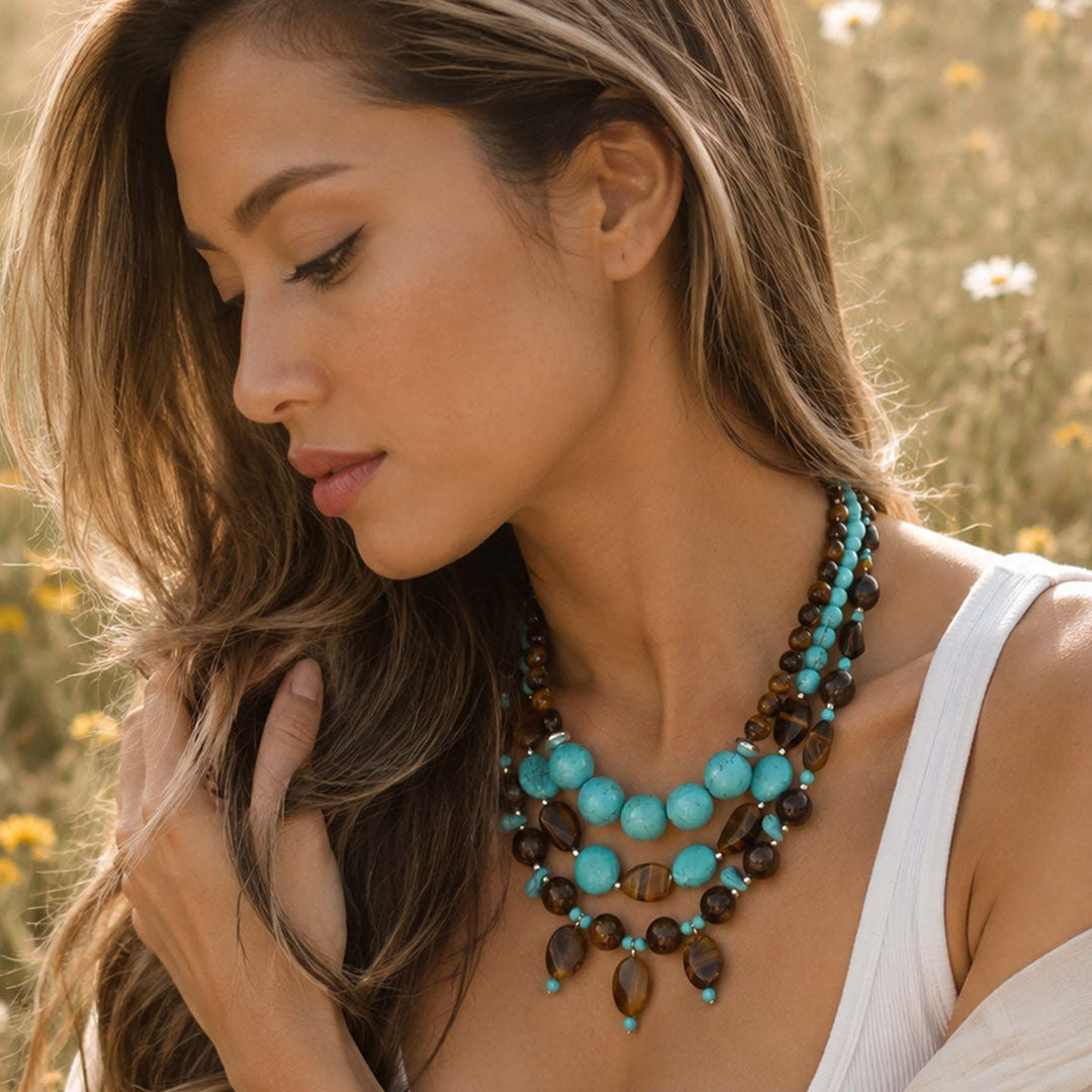 A woman wearing a multi-strand statement necklace with turquoise and dark brown beads, photographed outdoors in a sunlit field.
