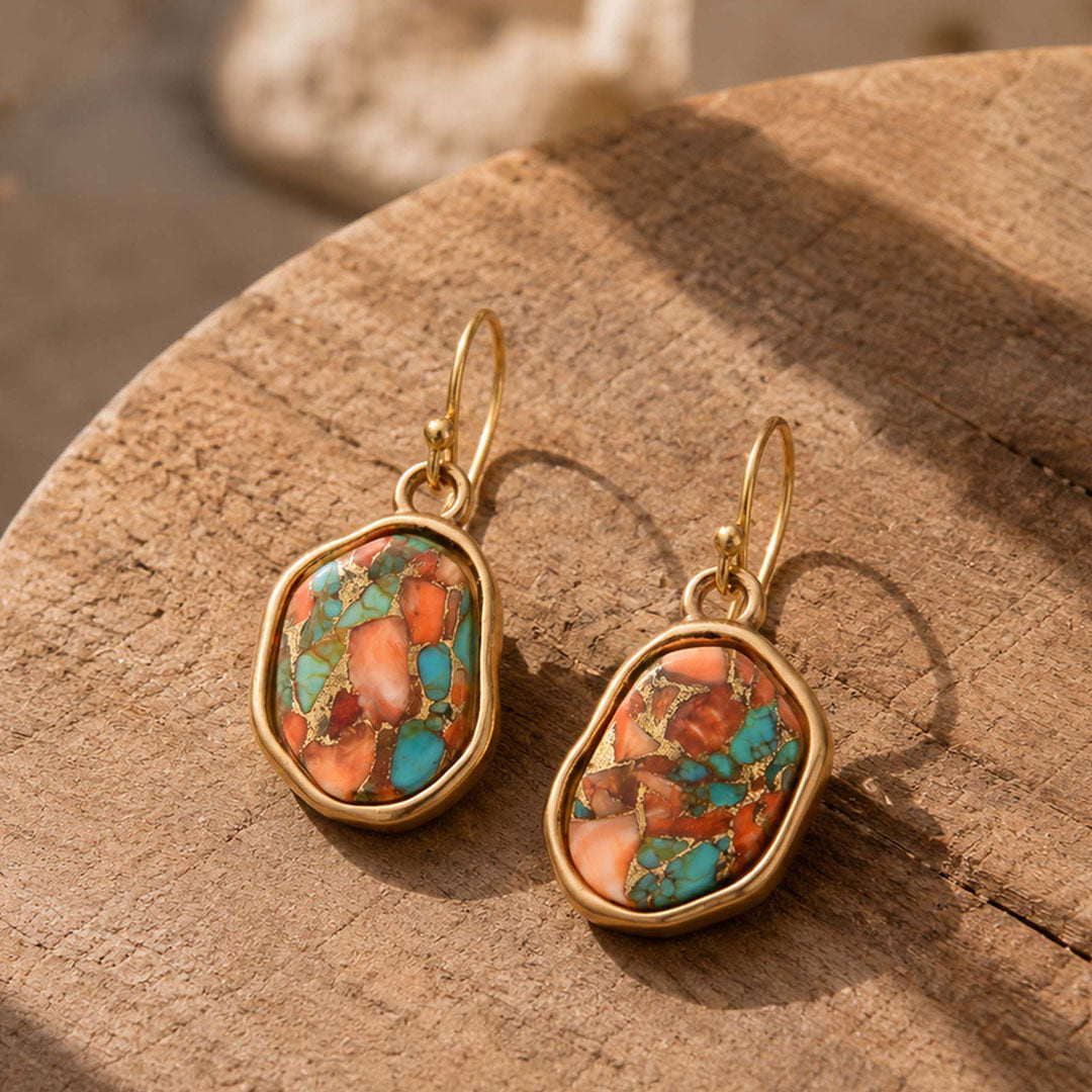 A pair of gold-tone drop earrings with irregular turquoise and coral stone inlays, displayed on a textured wooden surface.