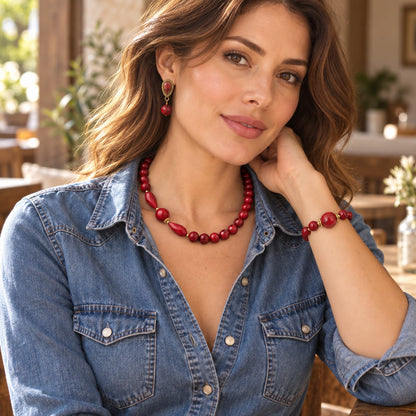 A woman wearing a red beaded necklace, matching red drop earrings, and a red beaded bracelet, styled with a blue denim shirt in a warm, softly blurred indoor setting.