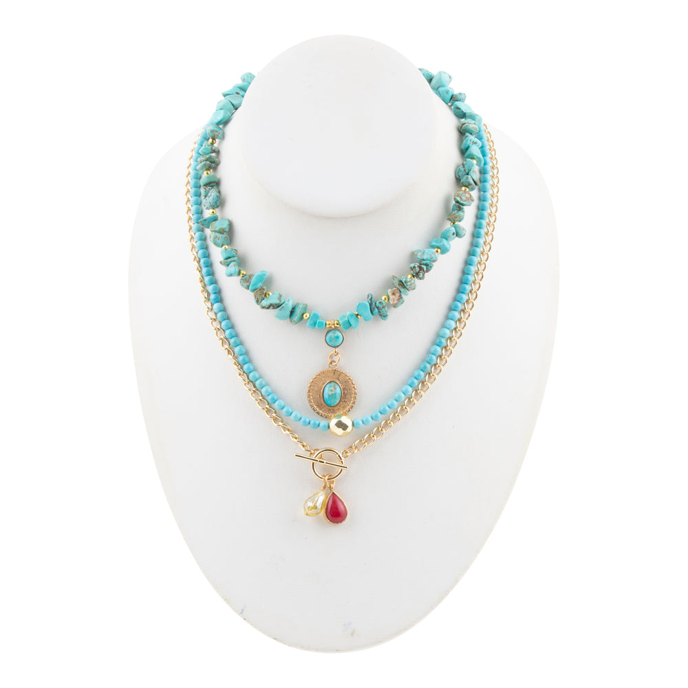A multi-strand bohemian necklace displayed on a white mannequin bust. The necklace features light blue beads, a gold chain, a turquoise stone pendant, and a dangling red teardrop charm.