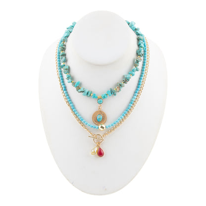 A multi-strand bohemian necklace displayed on a white mannequin bust. The necklace features light blue beads, a gold chain, a turquoise stone pendant, and a dangling red teardrop charm.