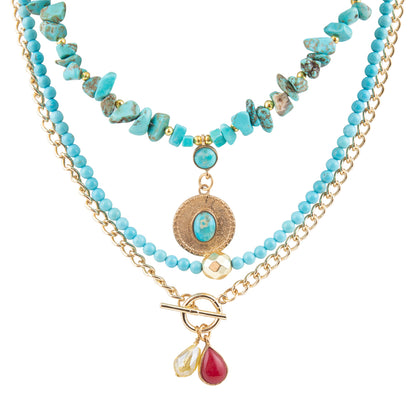 A multi-strand necklace featuring light blue turquoise beads, gold-tone chains, a textured round medallion with a turquoise center, and two dangling teardrop charms (one clear, one red) on a gold toggle clasp, shown against a plain white background.