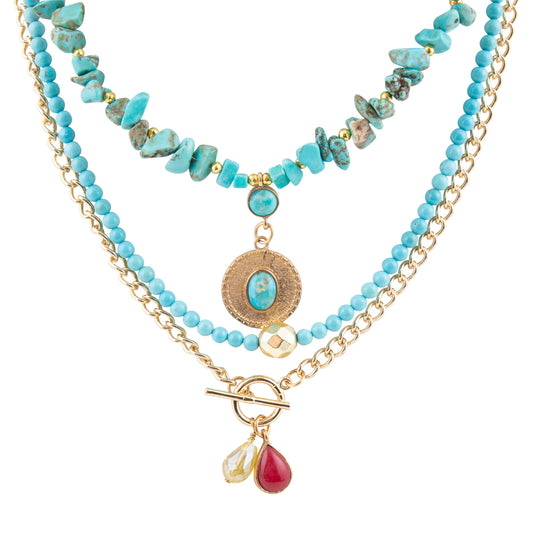 A multi-strand necklace featuring light blue turquoise beads, gold-tone chains, a textured round medallion with a turquoise center, and two dangling teardrop charms (one clear, one red) on a gold toggle clasp, shown against a plain white background.