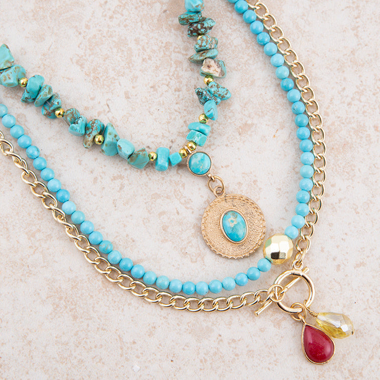 A multi-strand bohemian-style necklace featuring light blue turquoise beads, chunky turquoise stone beads, gold-tone chains, a round medallion charm, and two dangling teardrop charms (one red, one yellow) on a textured beige background.