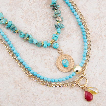 A multi-strand bohemian-style necklace featuring light blue turquoise beads, chunky turquoise stone beads, gold-tone chains, a round medallion charm, and two dangling teardrop charms (one red, one yellow) on a textured beige background.
