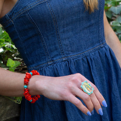 Person wearing a blue dress with a red bracelet and colorful ring, standing against a natural background.