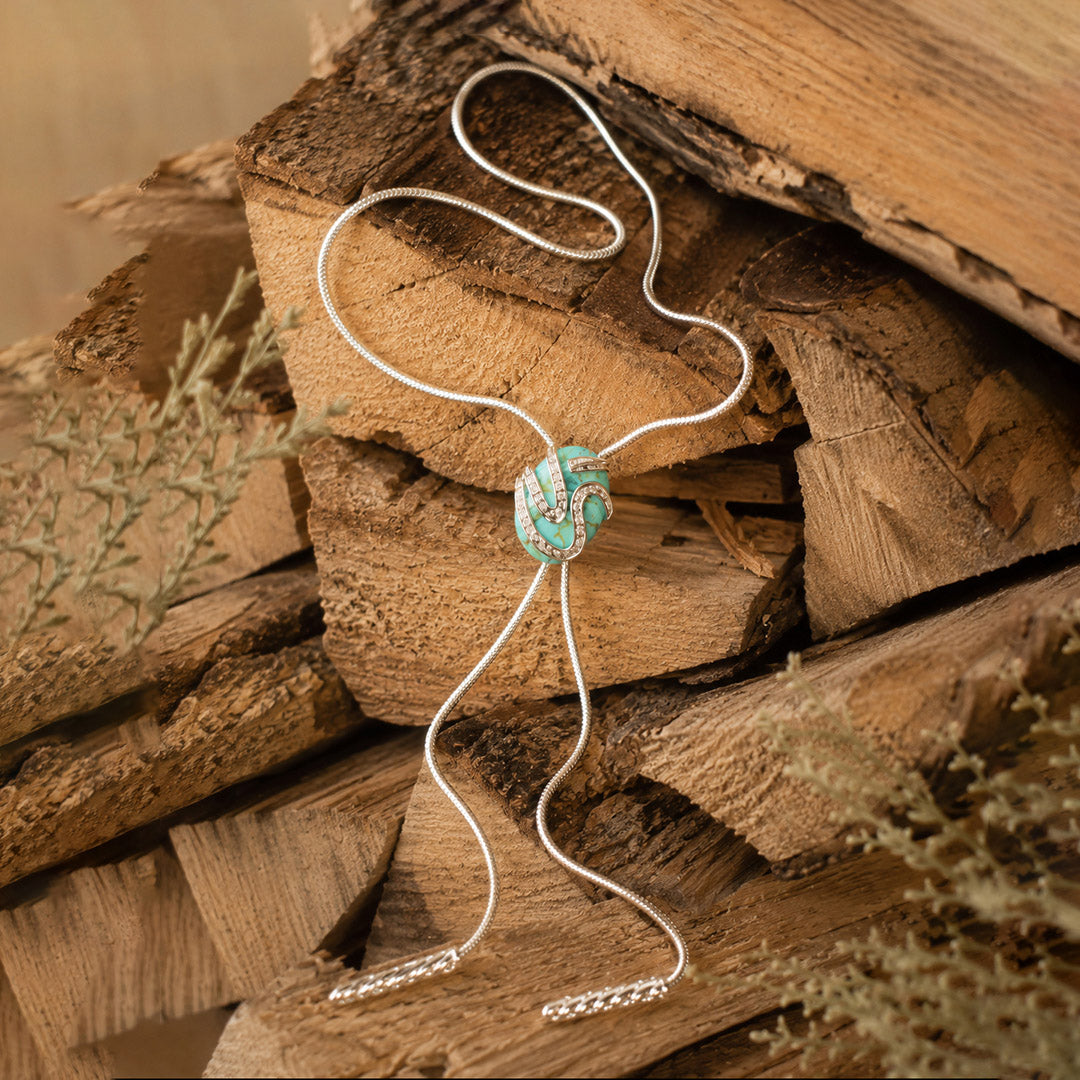 A silver-toned bolo tie necklace with a turquoise and white patterned center bead, displayed on rustic stacked wood logs.