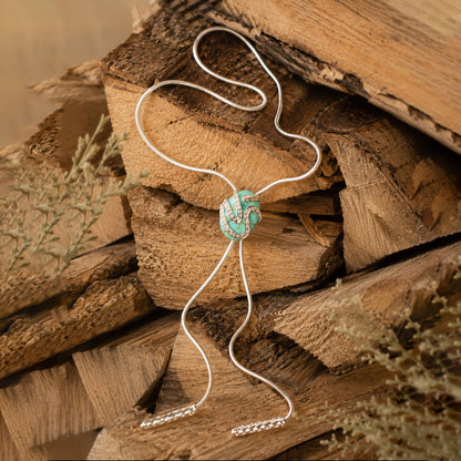 A silver-toned bolo tie necklace with a turquoise and white patterned center bead, displayed on rustic stacked wood logs.