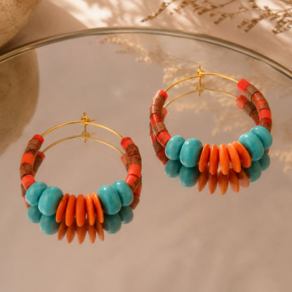 A pair of gold hoop earrings decorated with turquoise blue beads, orange disc beads, and brown patterned beads, displayed on a reflective glass surface.