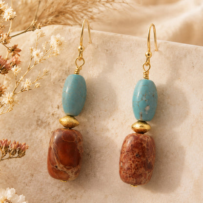 A pair of bohemian dangle earrings featuring light blue turquoise beads, brown stone beads, and gold-toned metal hooks, displayed on a textured beige stone surface with dried flowers.