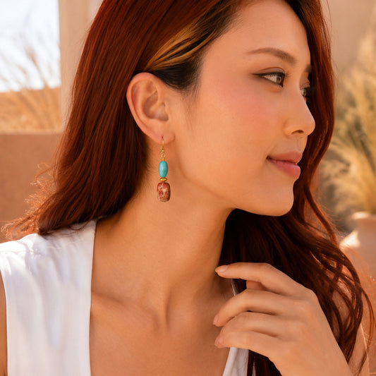 A woman in a white top wearing a dangling earring with a turquoise blue bead and a small gold-toned skull charm, photographed in warm natural light.