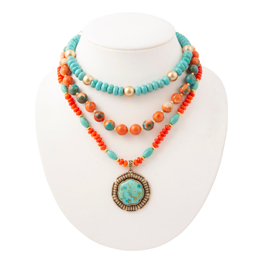 A multi-strand bohemian necklace displayed on a white mannequin bust. The necklace features turquoise beads, orange coral beads, small gold-toned pearls, and a large oval turquoise pendant with a bronze textured border.