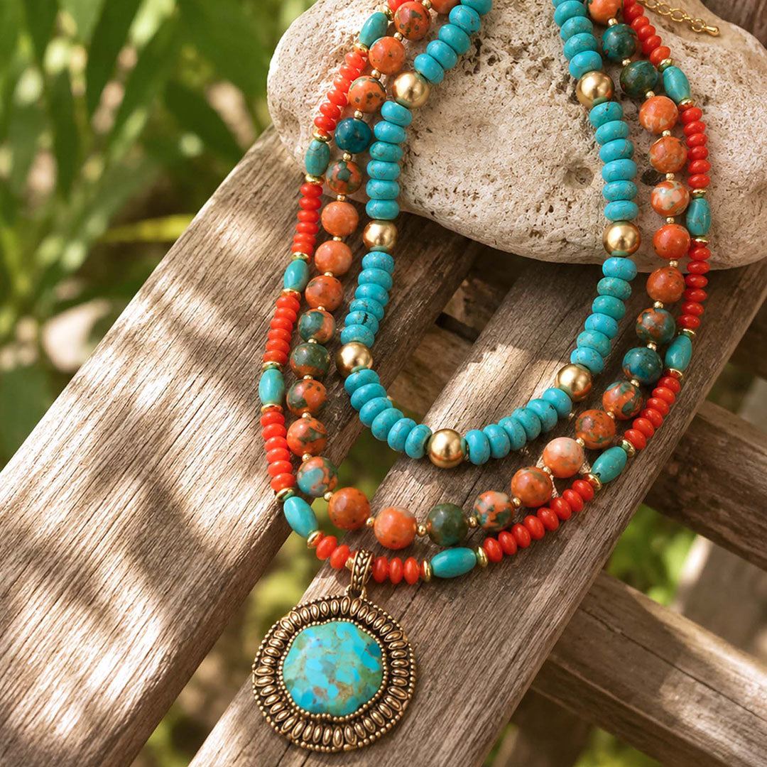 A bohemian-style multi-strand beaded necklace with turquoise, coral, and gold beads, featuring a large oval turquoise pendant, draped over a rustic wooden bench with a stone in the background.