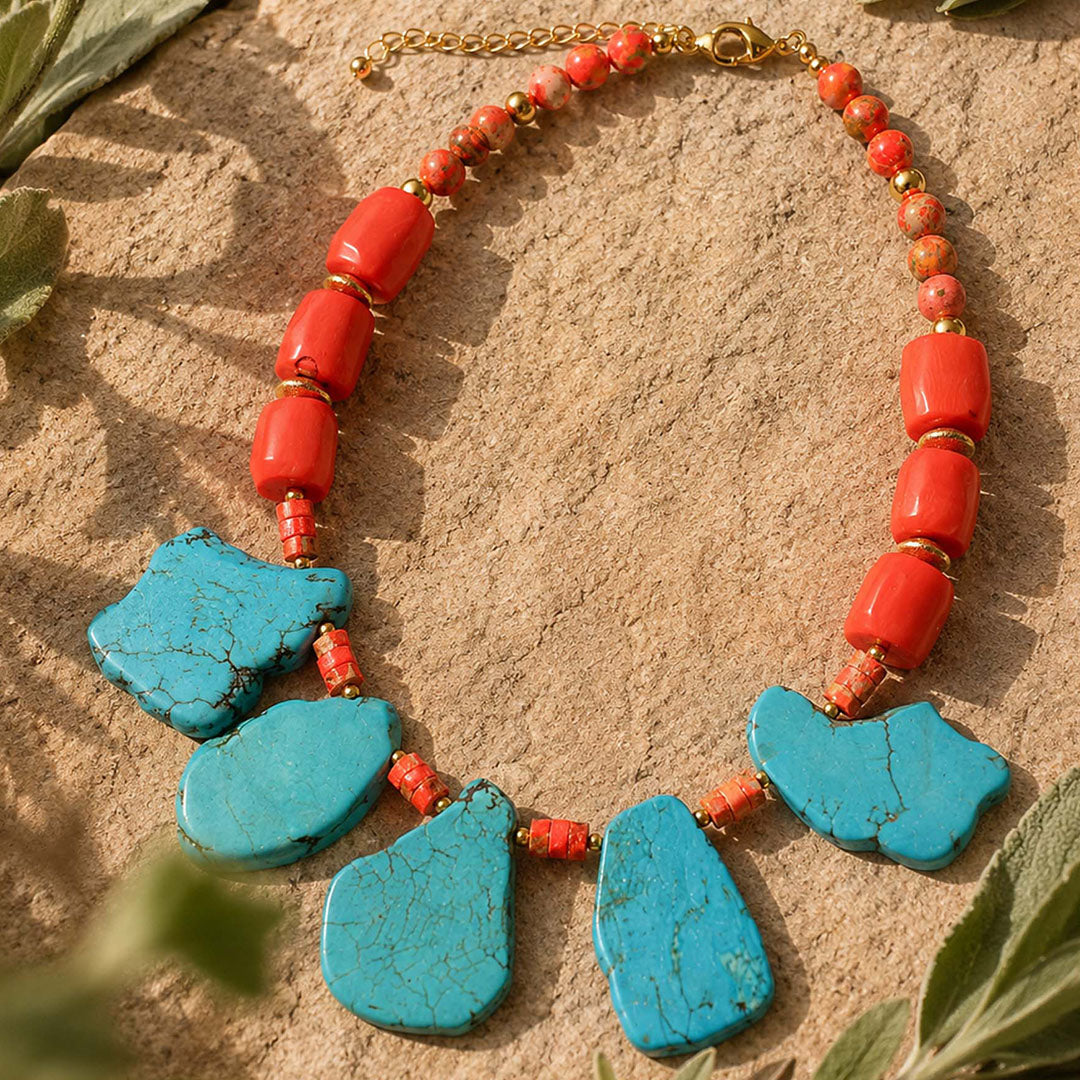 A statement necklace featuring five large turquoise-blue stone beads and multiple red-orange cylindrical beads separated by small gold spacers, displayed on a sandy surface with green leaves in the foreground.
