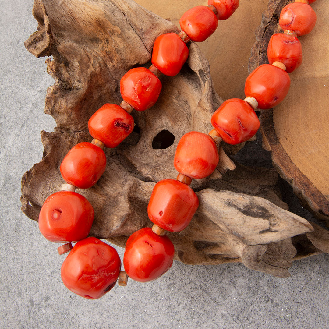 A beaded necklace made of glossy, rounded red coral-like beads with small light wood spacers, draped over a piece of natural driftwood on a gray surface.