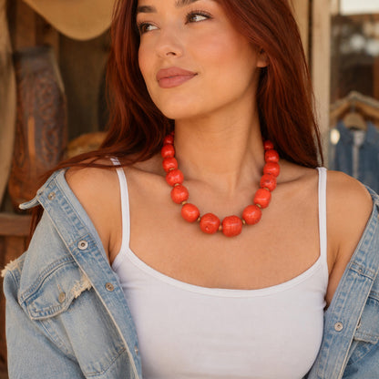 A woman with long auburn hair models a chunky coral-orange beaded necklace, styled with a white tank top and an open light-wash denim jacket in a rustic, warm-toned setting.