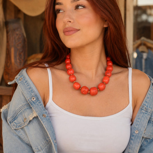 A woman with long auburn hair models a chunky coral-orange beaded necklace, styled with a white tank top and an open light-wash denim jacket in a rustic, warm-toned setting.