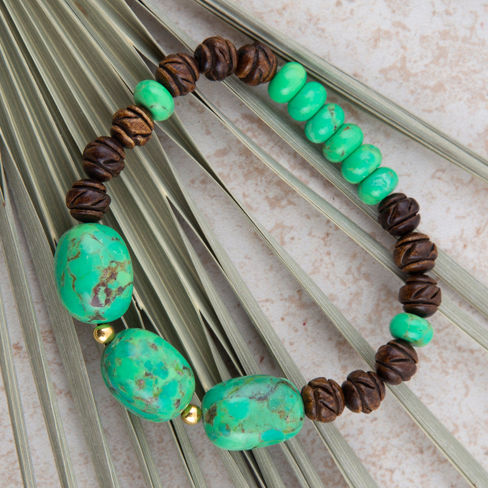 A beaded stretch bracelet featuring large turquoise-green marbled beads, smaller smooth green beads, dark brown textured wooden beads, and tiny gold spacer beads, displayed on a background of dried palm leaves.