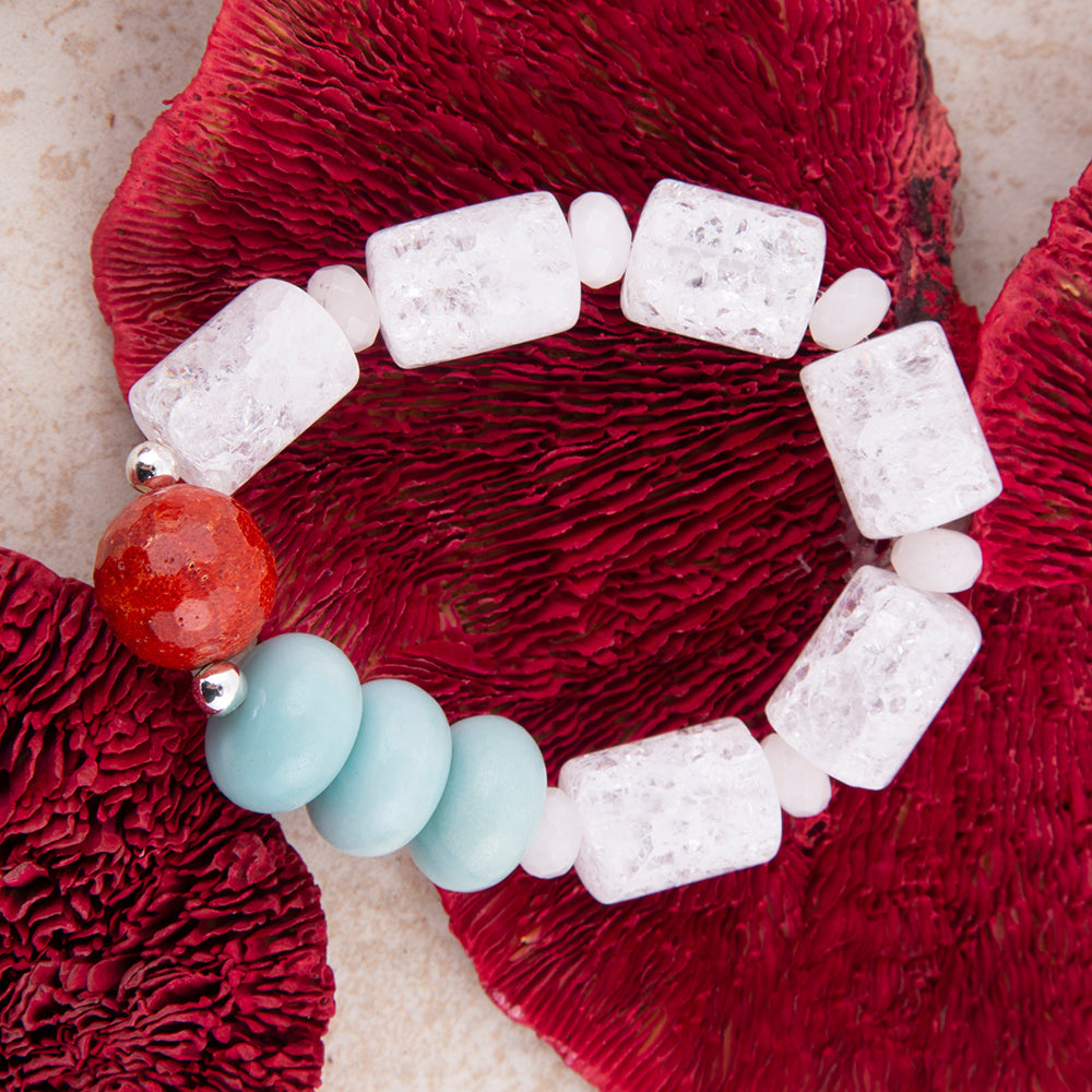 A beaded bracelet featuring textured white rectangular stones, four light blue round beads, a red-orange round bead, and small silver spacer beads, displayed on a textured red mushroom cap over a beige surface.