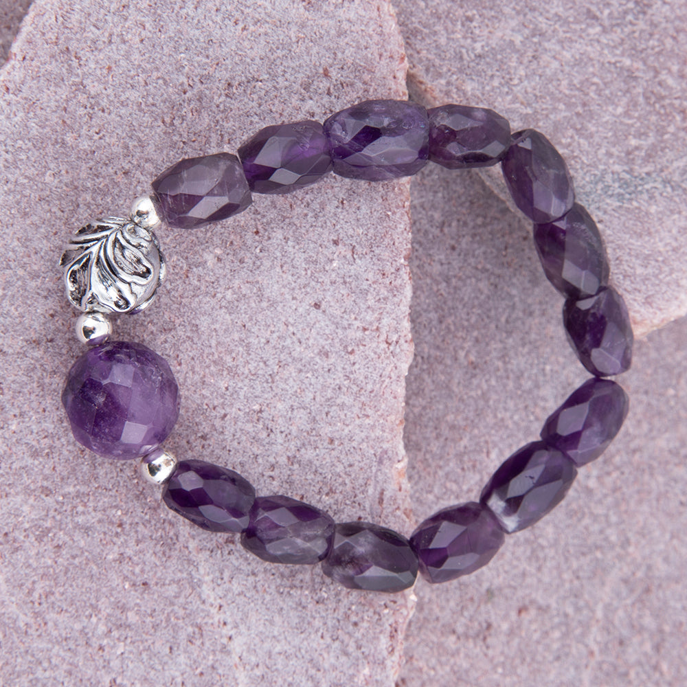 A beaded stretch bracelet made of faceted purple gemstone beads and a large polished purple bead, accented by silver-toned spacer beads and a decorative silver filigree bead, displayed on a textured pinkish stone surface.