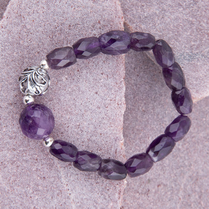A beaded stretch bracelet made of faceted purple gemstone beads and a large polished purple bead, accented by silver-toned spacer beads and a decorative silver filigree bead, displayed on a textured pinkish stone surface.