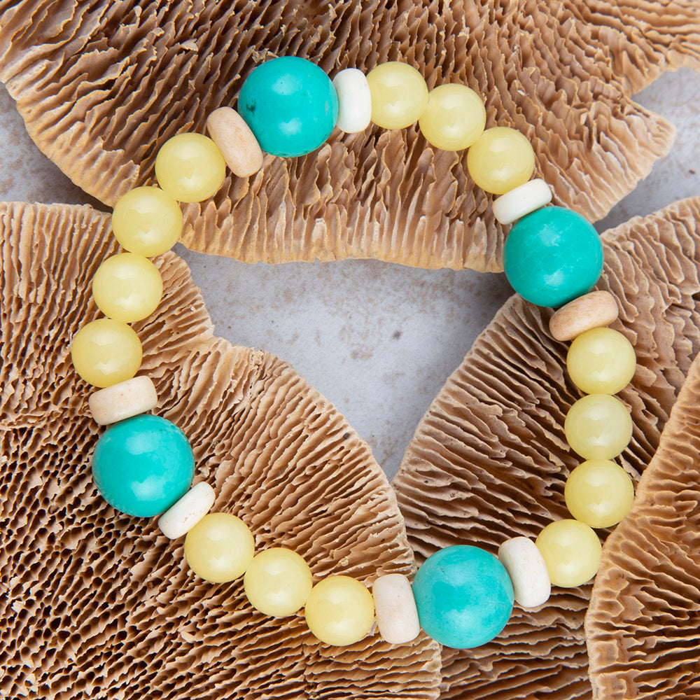 A beaded bracelet made of turquoise-blue and pale yellow round beads, accented with small natural wood beads, arranged in a circle over brown mushrooms on a light gray surface.