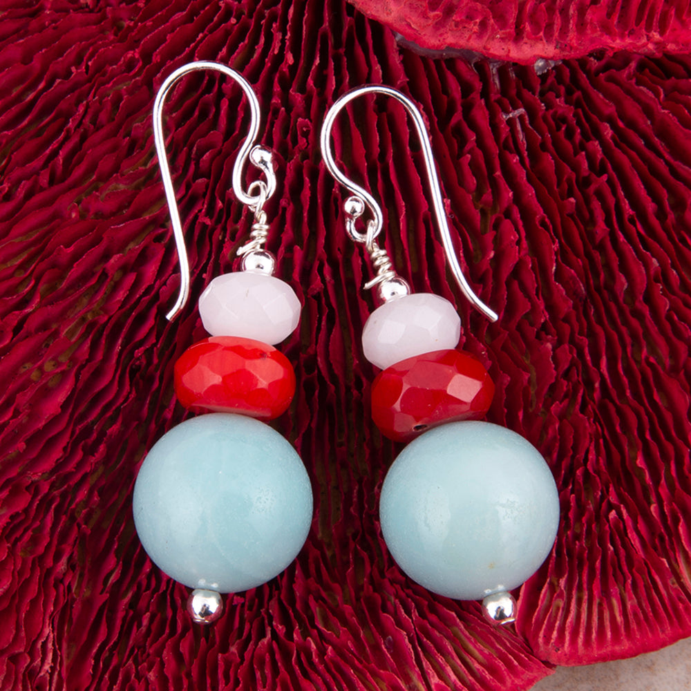 A pair of dangling earrings with silver fishhook wires, featuring a stack of beads: a pale pink stone, a faceted red stone, and a large light blue round bead, displayed on a textured dark red surface.